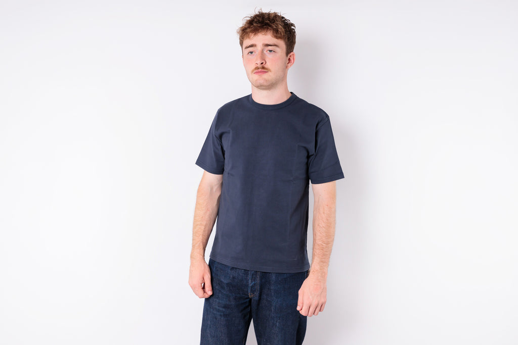 A man wearing a dark navy blue crewneck t-shirt and dark blue jeans, standing against a plain white studio background.