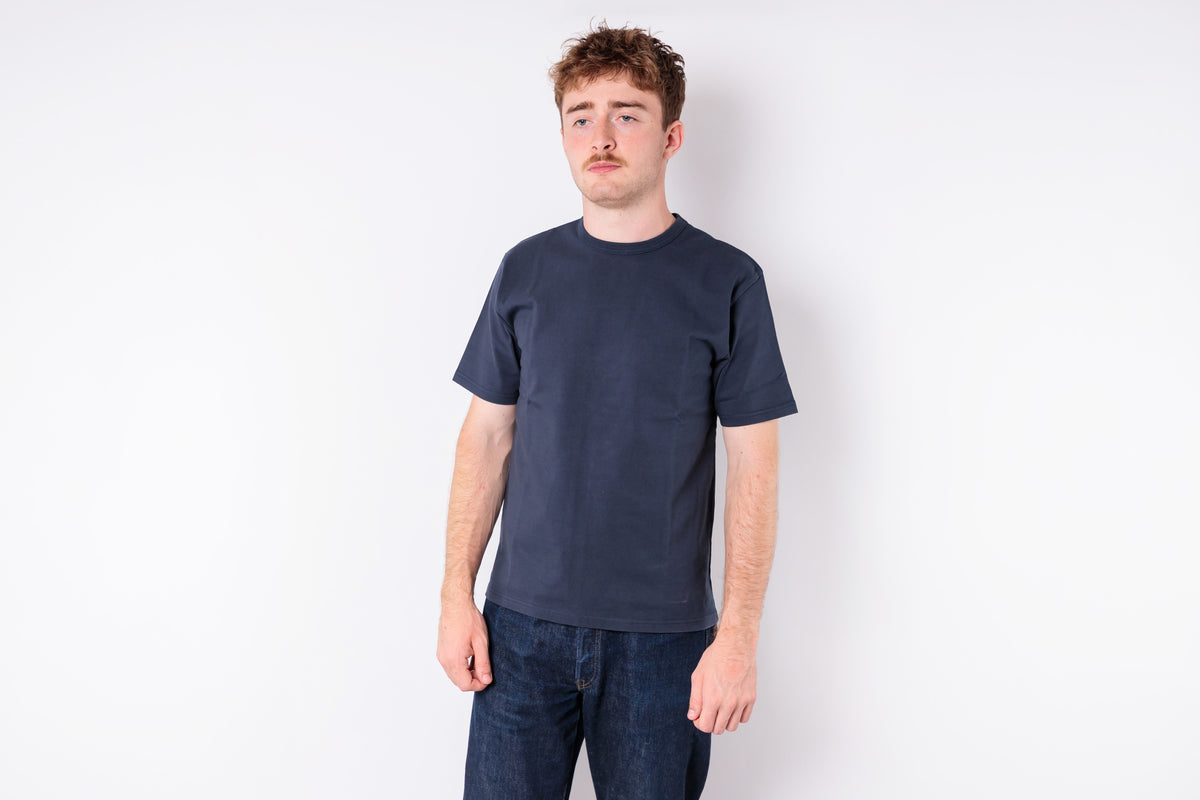 A man wearing a dark navy blue crewneck t-shirt and dark blue jeans, standing against a plain white studio background.