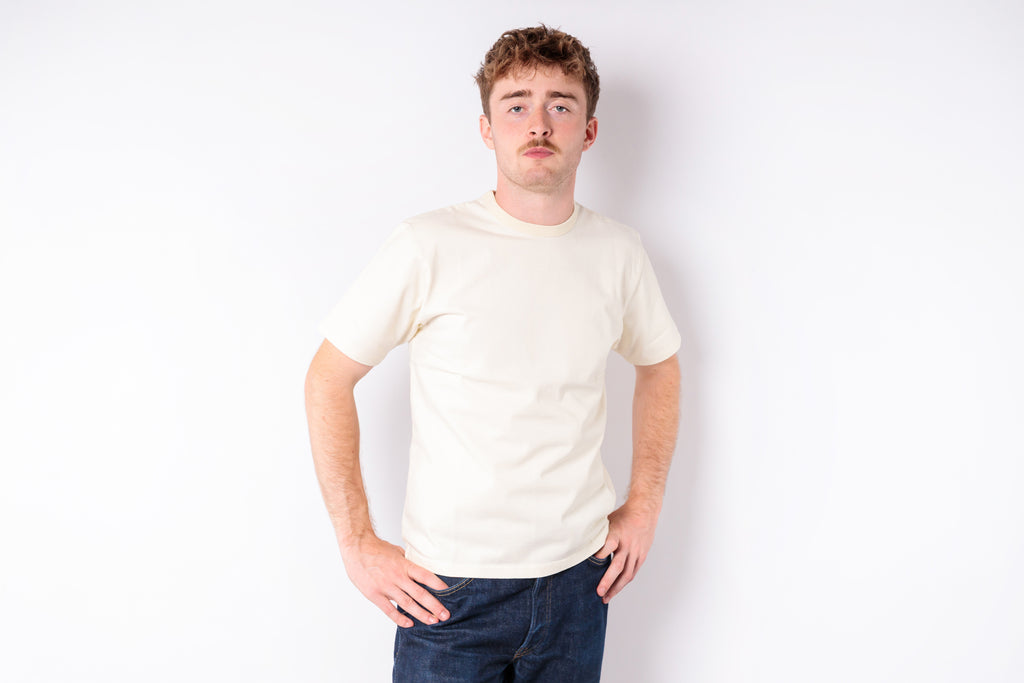 A man wearing a plain cream-colored short-sleeve t-shirt and dark blue jeans, standing against a solid white background.