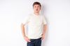 A man wearing a plain cream-colored short-sleeve t-shirt and dark blue jeans, standing against a solid white background.