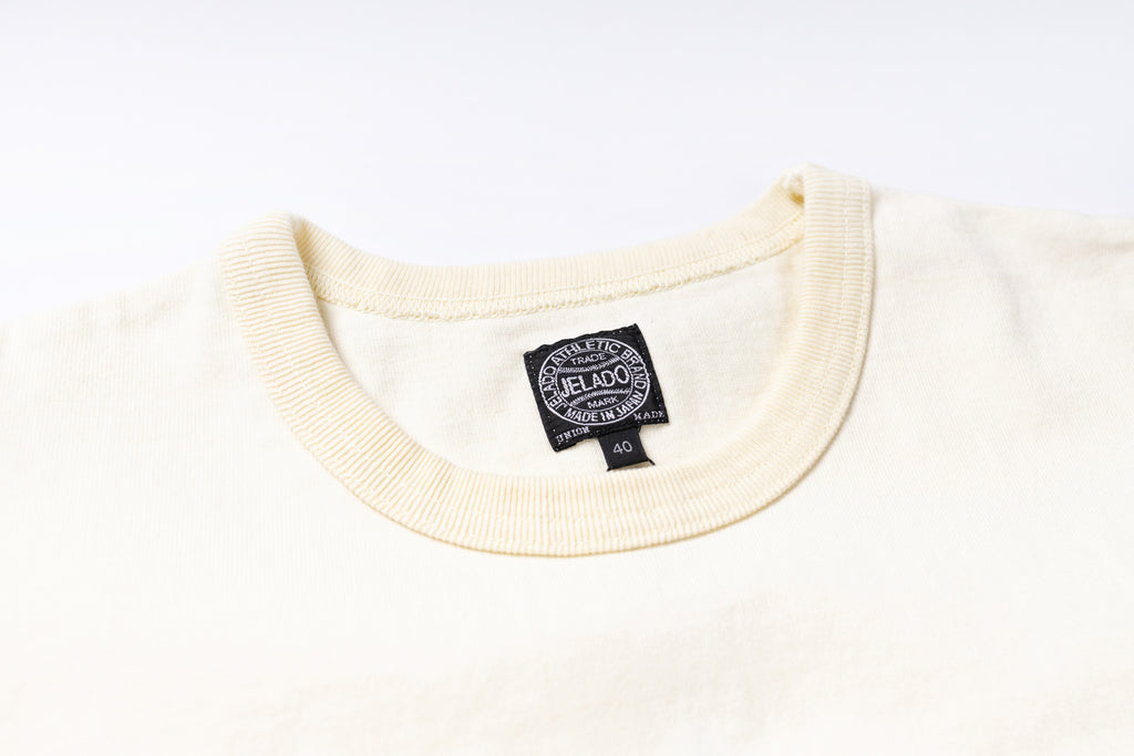 Close-up of a cream-colored knit crewneck sweater showing the ribbed collar and a black circular 'JELADO' brand tag inside the neckline.