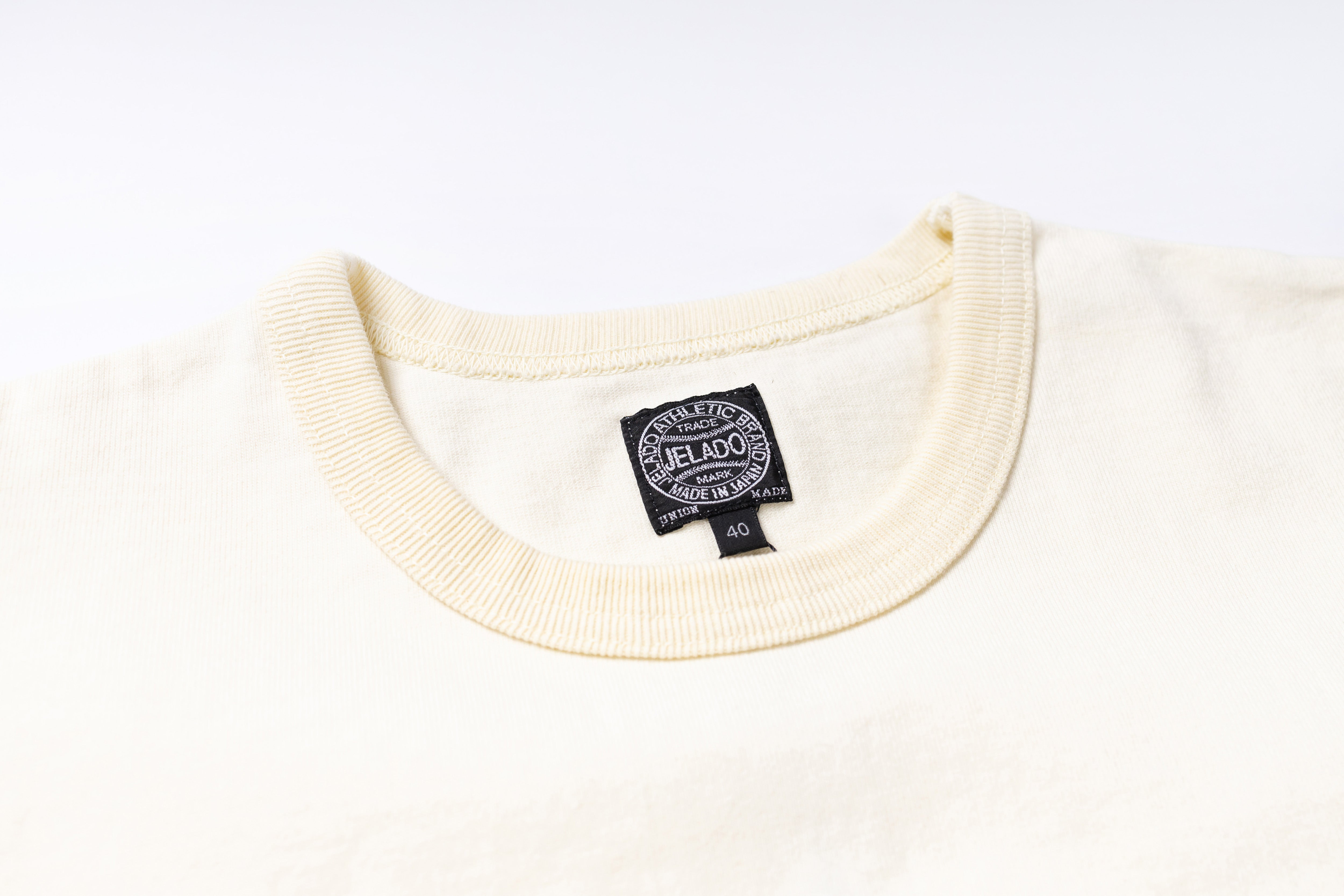 Close-up of a cream-colored knit crewneck sweater showing the ribbed collar and a black circular 'JELADO' brand tag inside the neckline.
