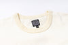 Close-up of a cream-colored knit crewneck sweater showing the ribbed collar and a black circular 'JELADO' brand tag inside the neckline.