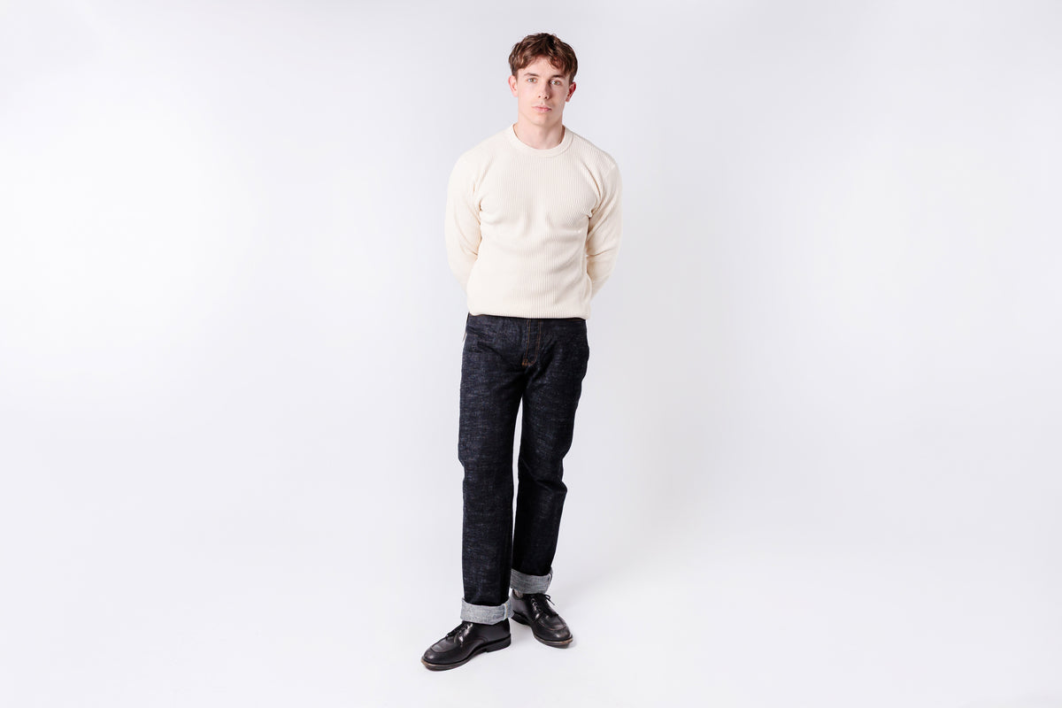 Man wearing a cream sweater and dark pants on a white background