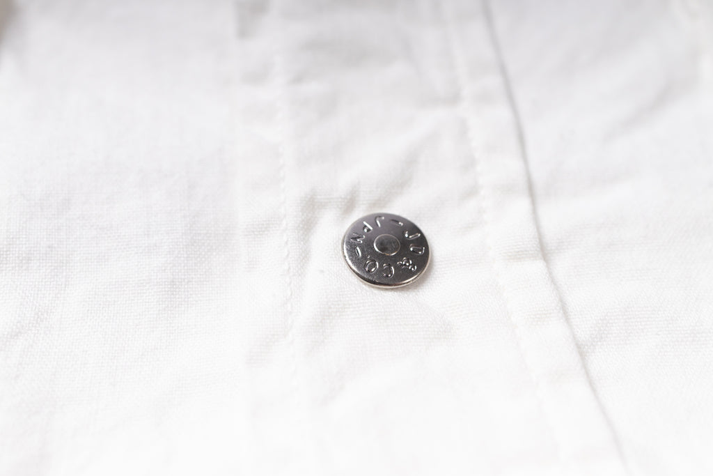 A close-up of a white textured fabric garment featuring a small, round, silver-toned metal button.