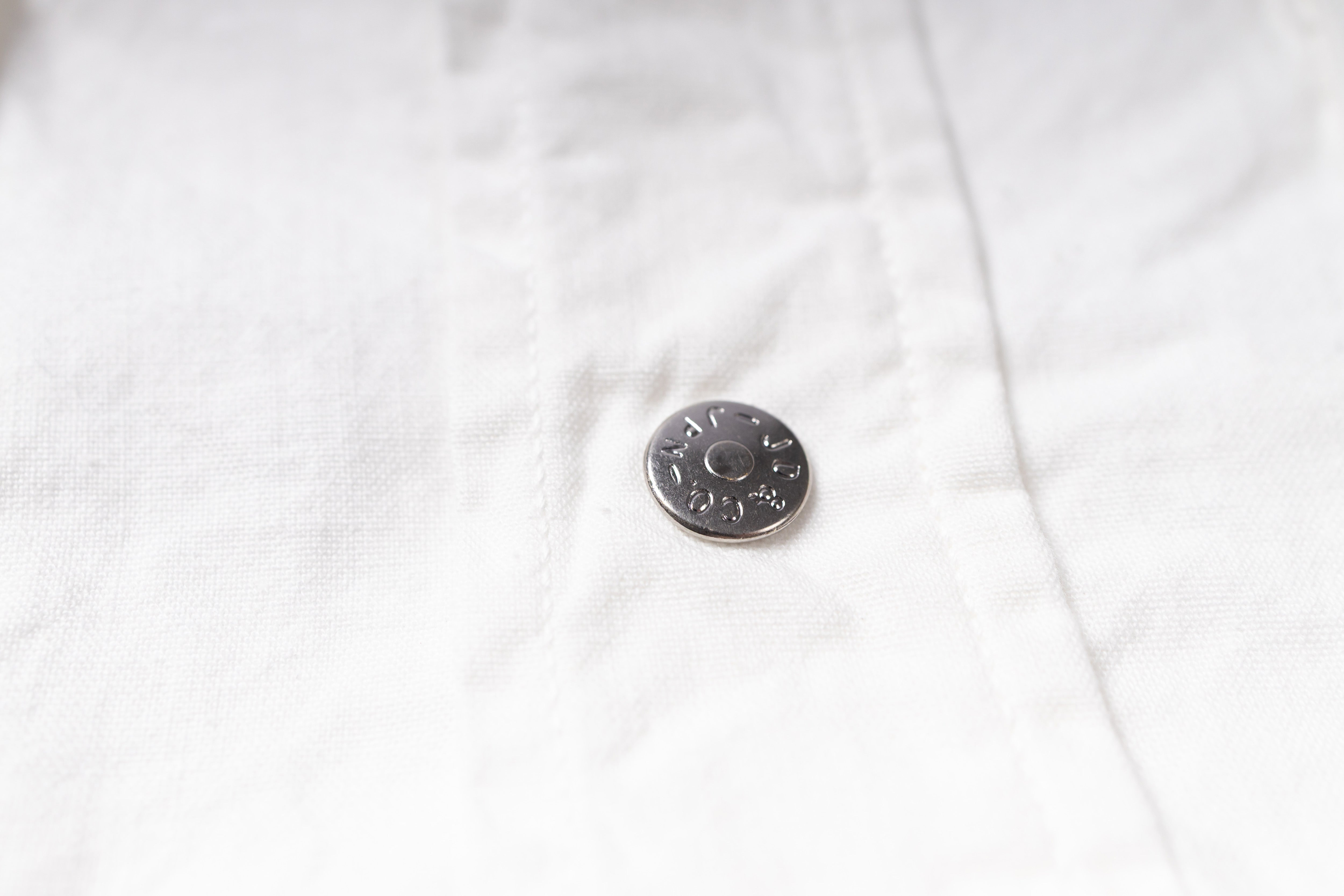 A close-up of a white textured fabric garment featuring a small, round, silver-toned metal button.
