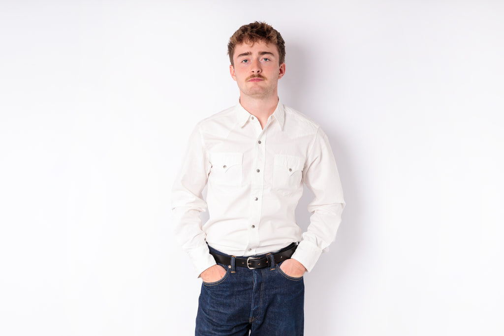A man with a mustache wearing a cream-colored long-sleeve button-down shirt and dark blue jeans, standing against a plain white background.