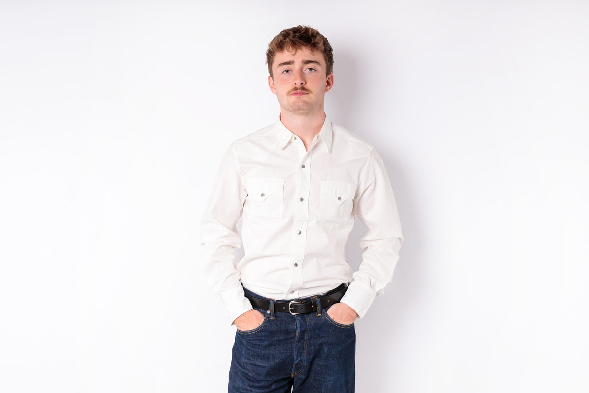 A man with a mustache wearing a cream-colored long-sleeve button-down shirt and dark blue jeans, standing against a plain white background.