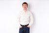 A man with a mustache wearing a cream-colored long-sleeve button-down shirt and dark blue jeans, standing against a plain white background.