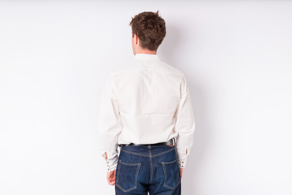 A man viewed from behind wearing a white long-sleeve button-up shirt and blue jeans against a plain white background.