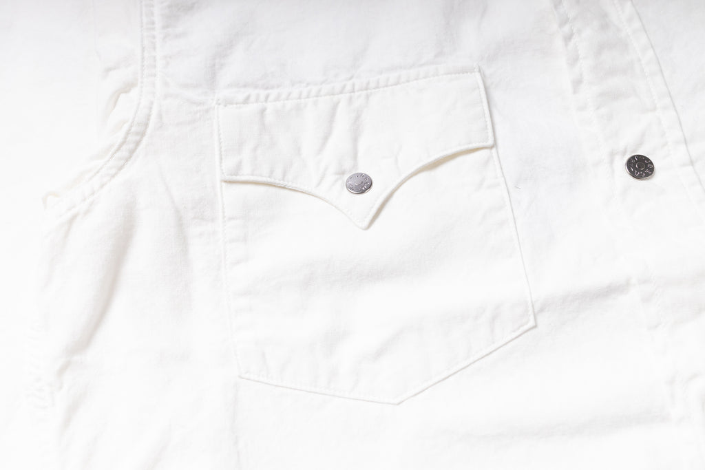 Close-up of a white denim garment showing a front chest pocket with a silver-toned button and part of a dark button on the right side.