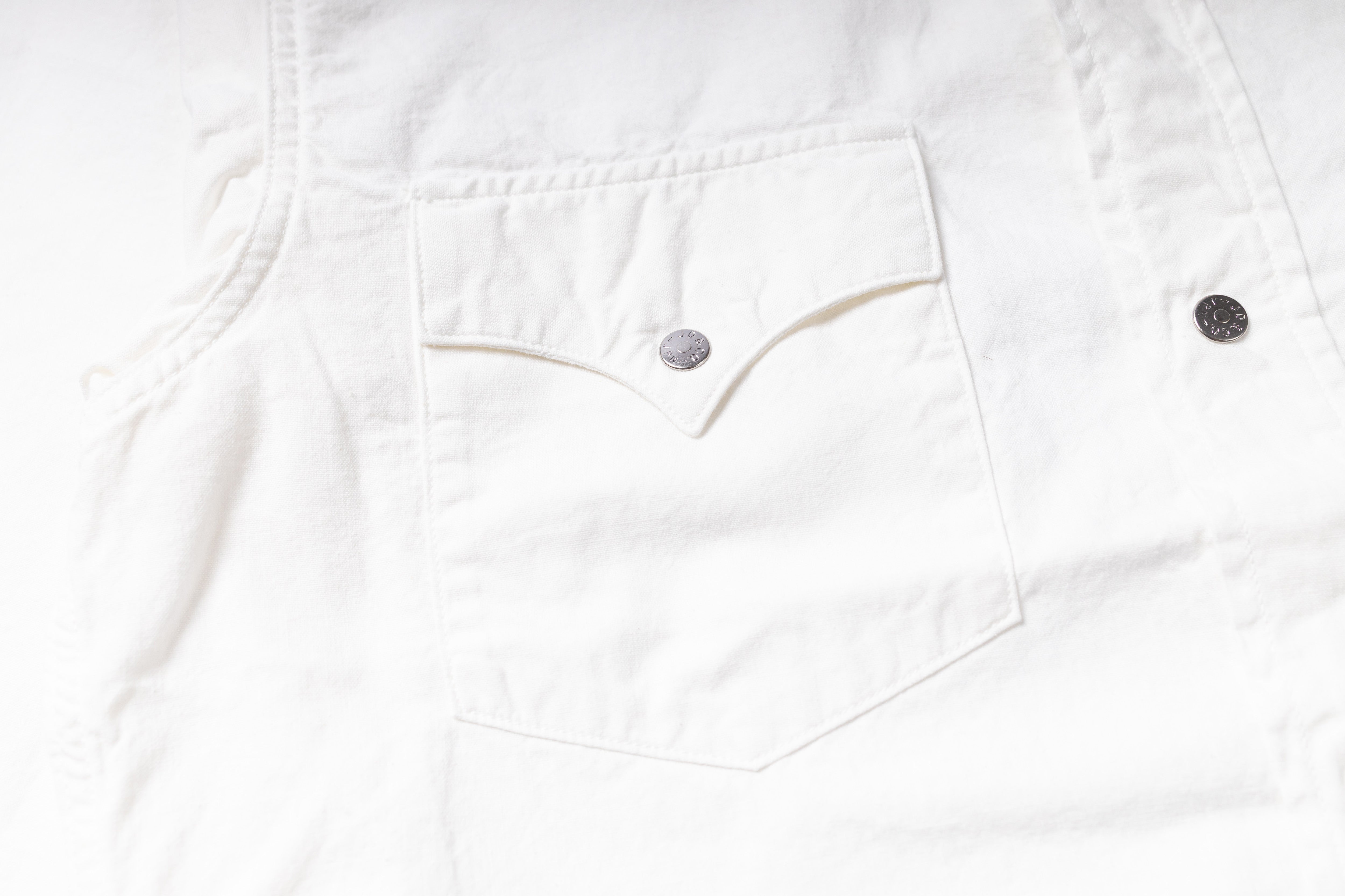 Close-up of a white denim garment showing a front chest pocket with a silver-toned button and part of a dark button on the right side.