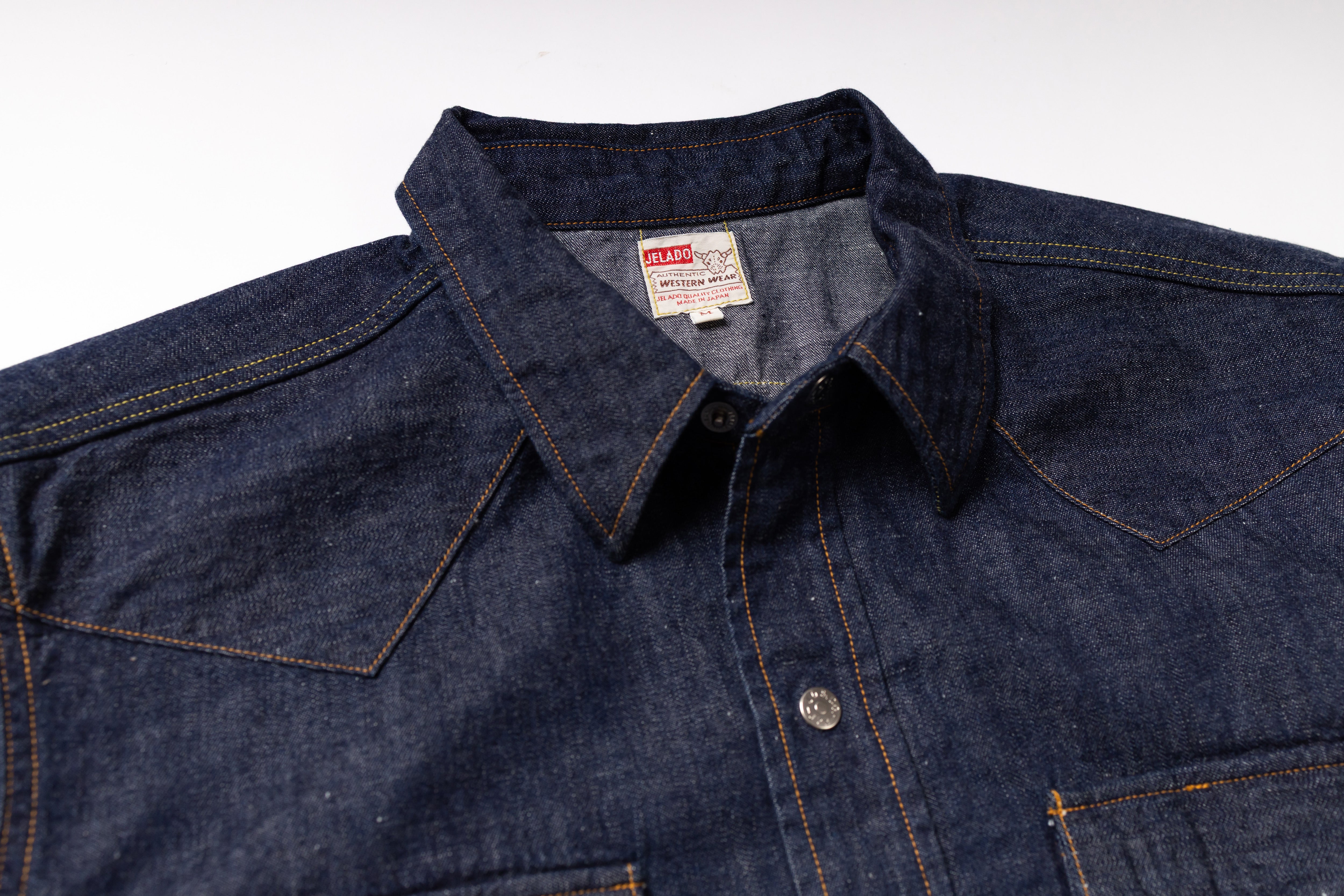 Close-up of a dark indigo denim shirt jacket with contrast orange stitching, a pointed collar, and a visible U.S.