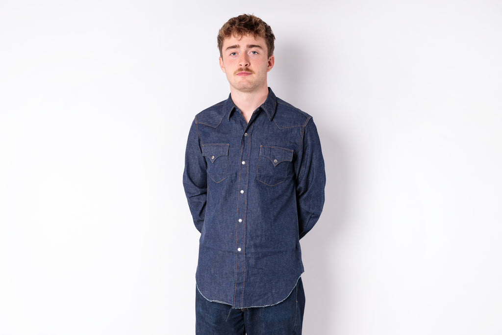 A man wearing a dark indigo denim long-sleeve western-style shirt with two buttoned chest pockets, standing against a plain white studio background.