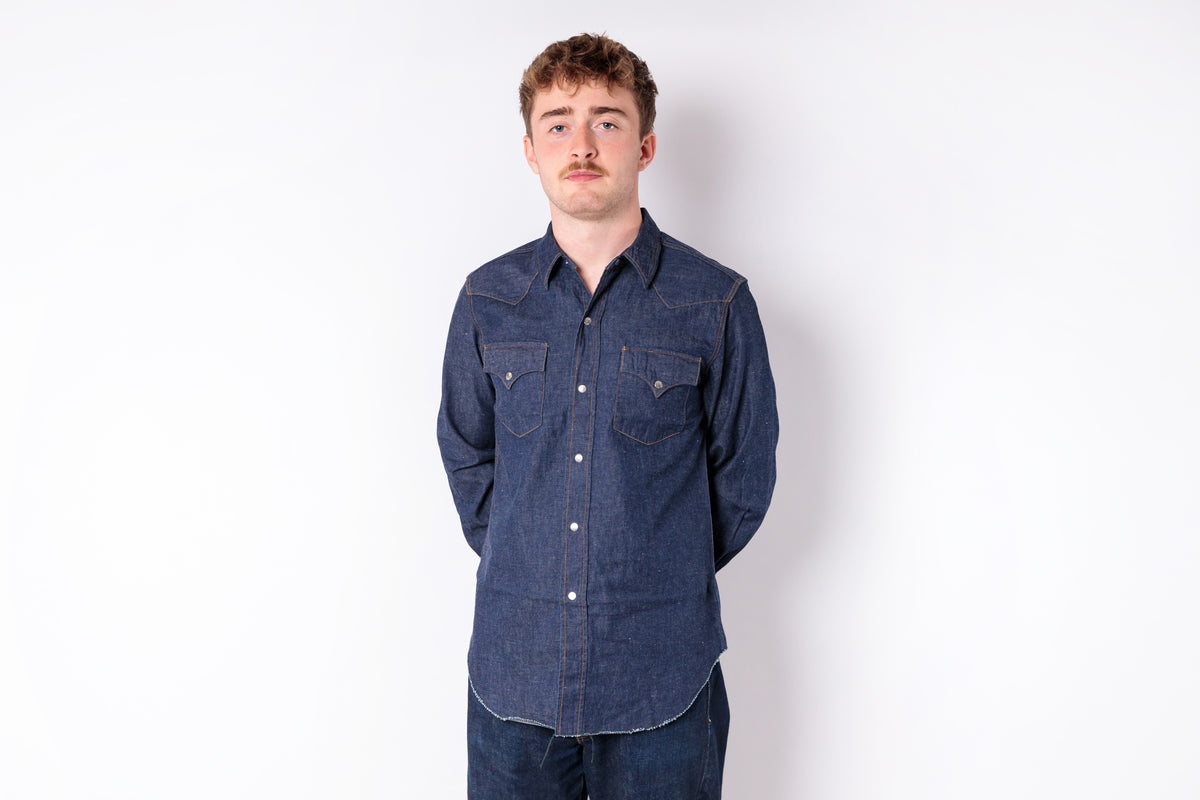 A man wearing a dark indigo denim long-sleeve western-style shirt with two buttoned chest pockets, standing against a plain white studio background.