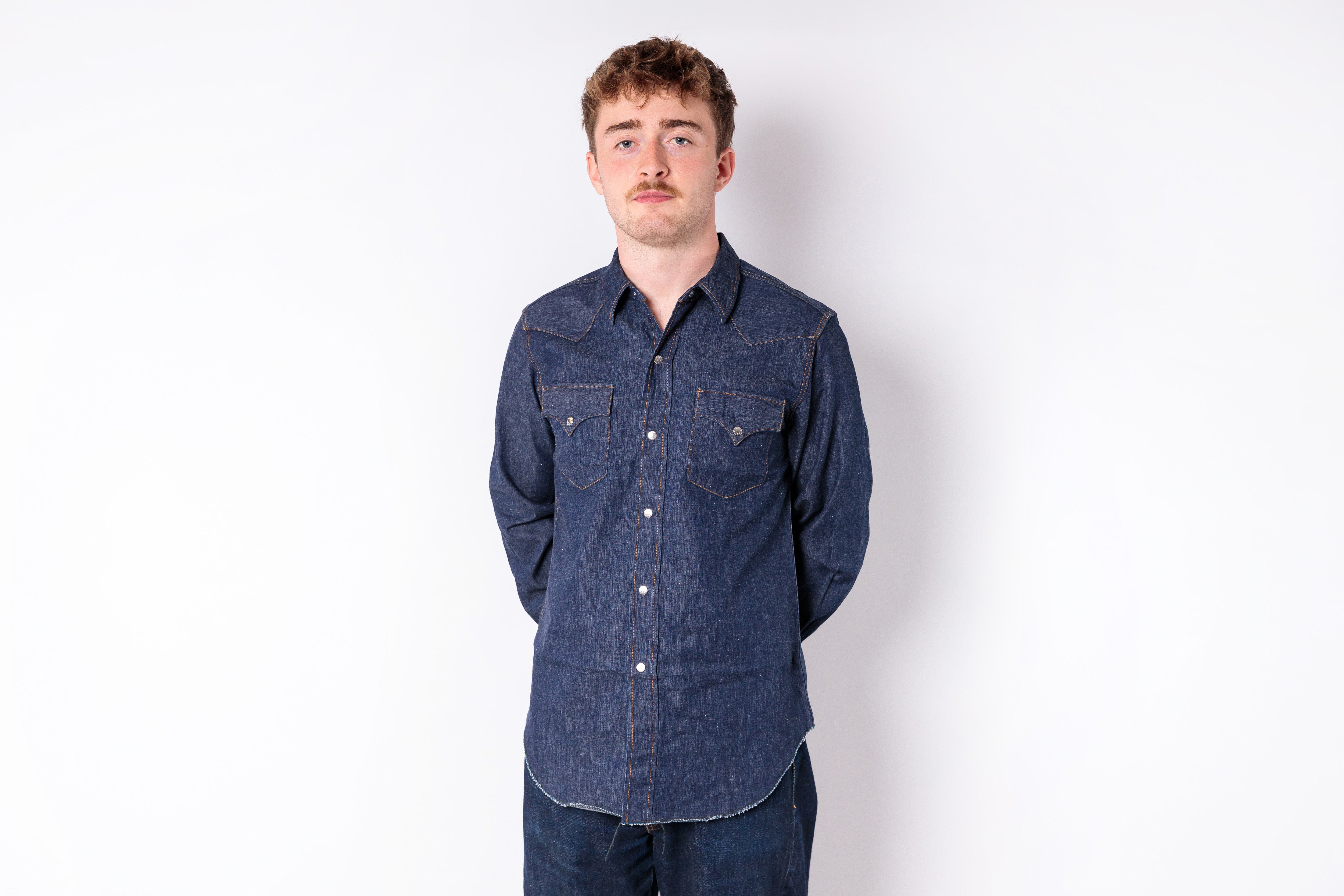A man wearing a dark indigo denim long-sleeve western-style shirt with two buttoned chest pockets, standing against a plain white studio background.