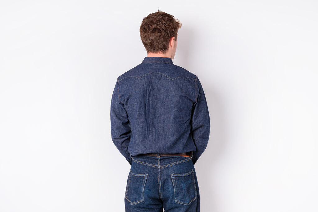 A man viewed from behind wearing a dark blue denim long-sleeve button-up shirt and matching blue jeans, standing against a plain white wall.