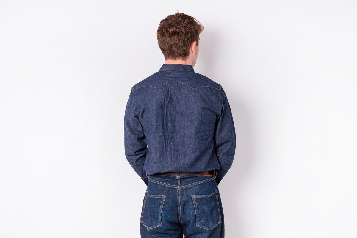 A man viewed from behind wearing a dark blue denim long-sleeve button-up shirt and matching blue jeans, standing against a plain white wall.