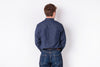 A man viewed from behind wearing a dark blue denim long-sleeve button-up shirt and matching blue jeans, standing against a plain white wall.