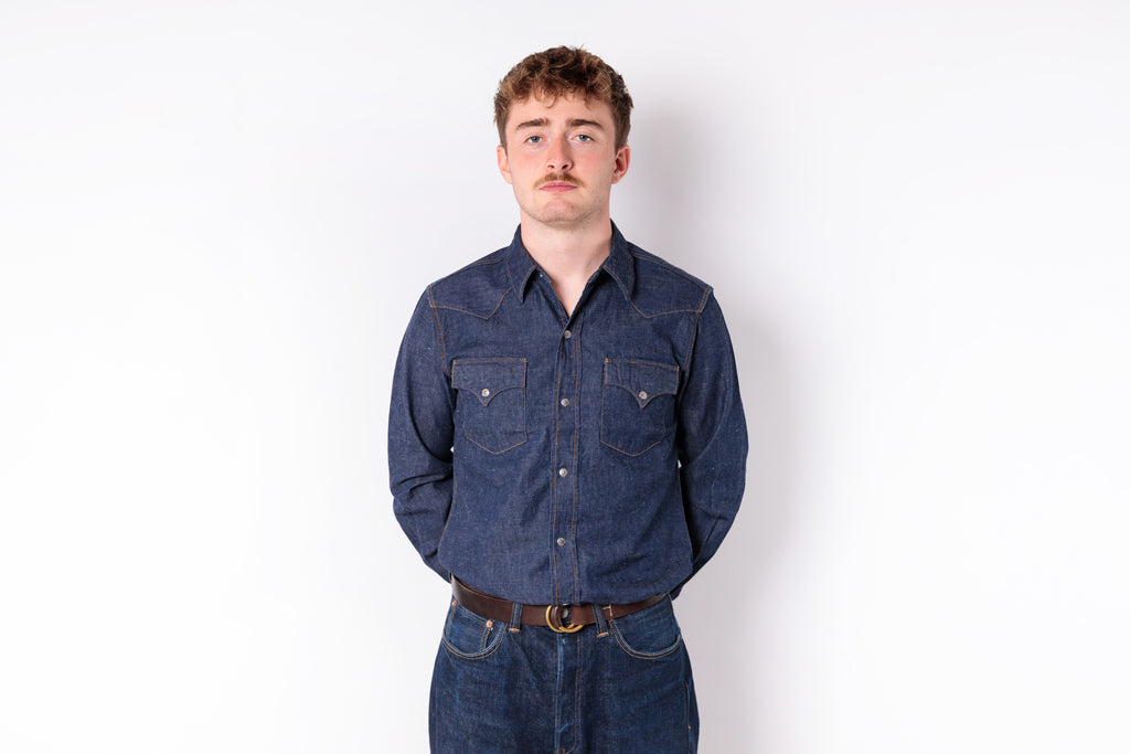 A man wearing a dark blue denim long-sleeve button-up shirt and matching blue jeans, standing against a plain white background.