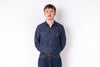 A man wearing a dark blue denim long-sleeve button-up shirt and matching blue jeans, standing against a plain white background.