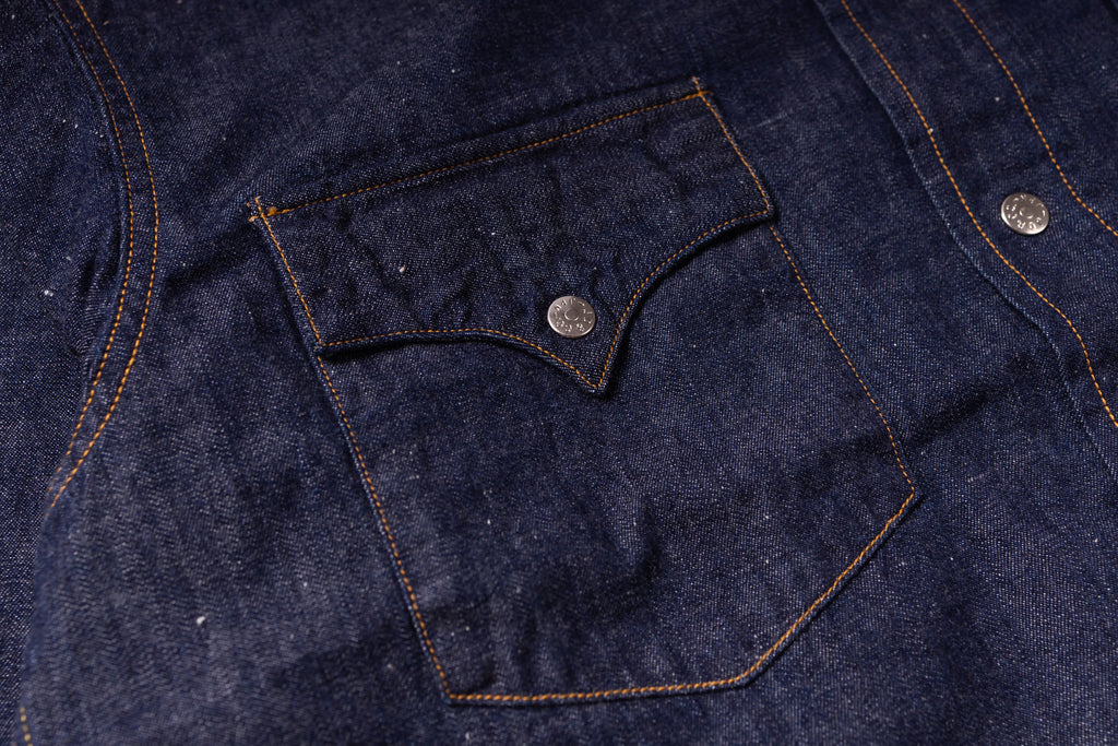 Close-up of a dark indigo denim garment showing a buttoned chest pocket with contrast orange stitching and a silver metal button.