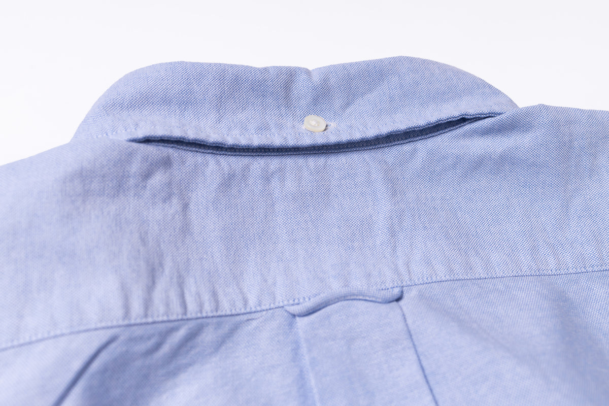 Close-up of the back collar of a light blue textured shirt, showing a single button and dark contrast stitching.
