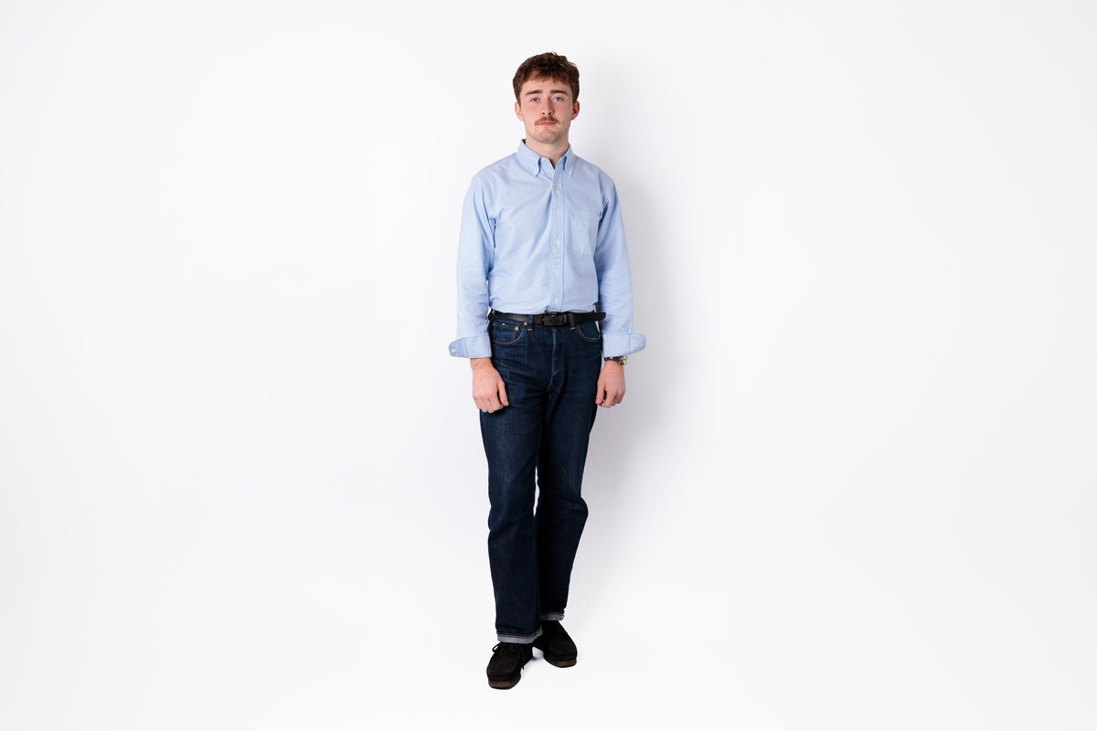 A man wearing a light blue long-sleeve button-down shirt, dark blue jeans, and black shoes, standing against a plain white background.