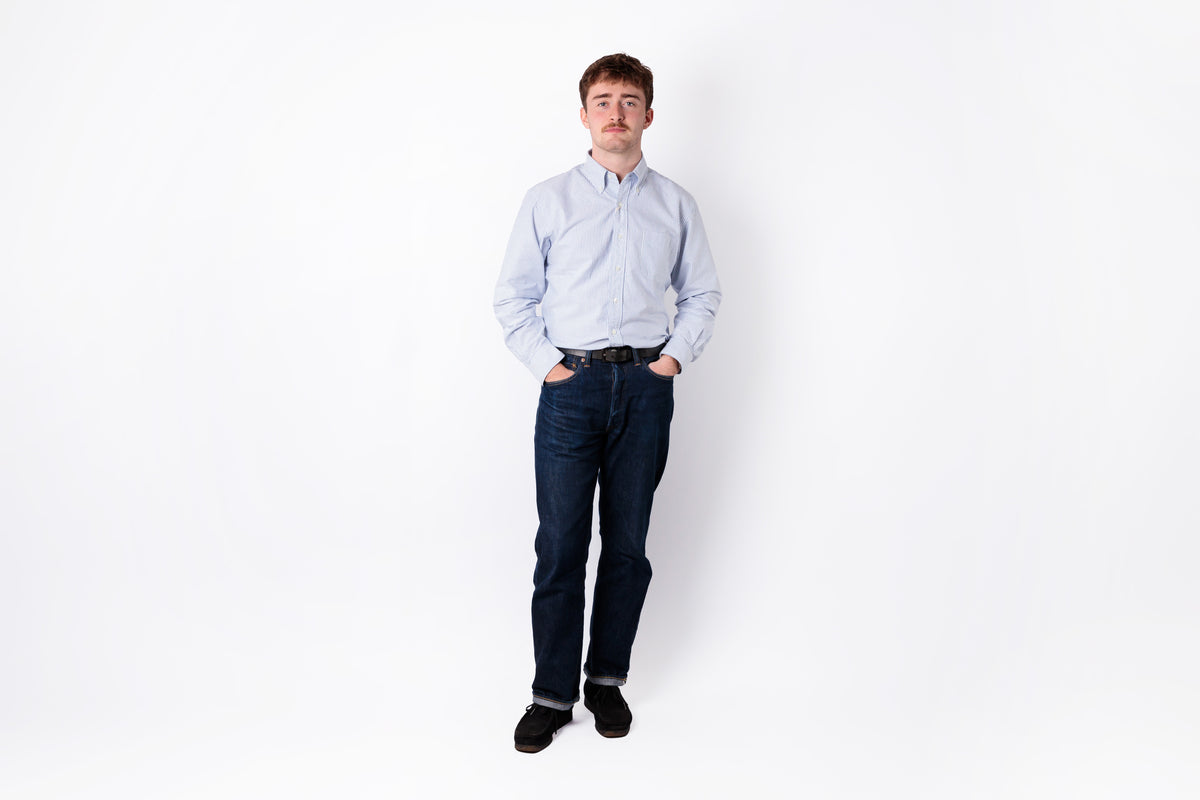 A man wearing a light blue and white striped button-down shirt, dark blue jeans, and black shoes, standing against a plain white background.