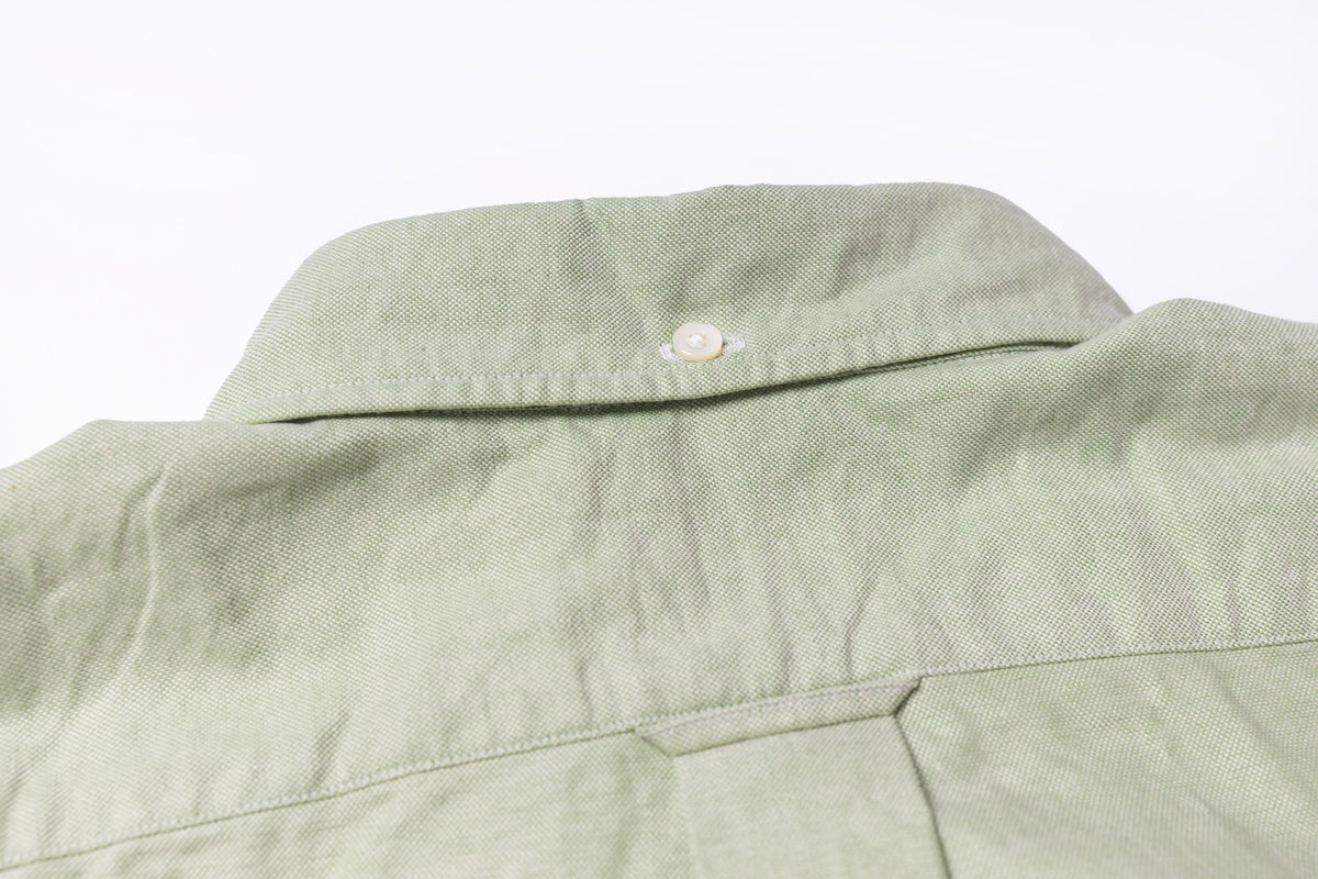 Close-up of the back collar and upper back of a light sage green linen shirt, showing a single white button and fabric texture against a white background.