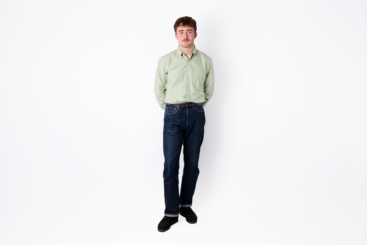 A man wearing a light sage green button-down shirt, dark blue jeans, and black shoes, standing against a plain white background.