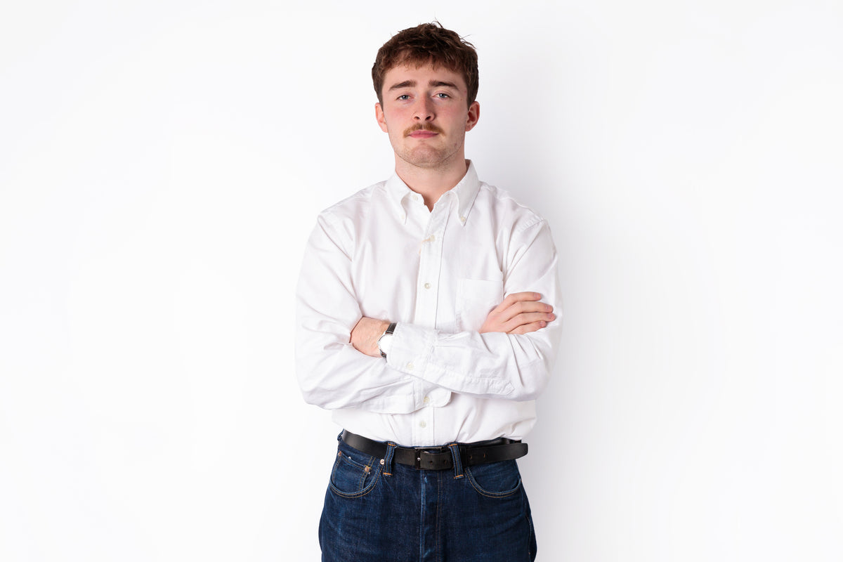 A young man with a mustache stands against a white wall with his arms crossed, wearing a white button-down shirt and dark blue jeans.