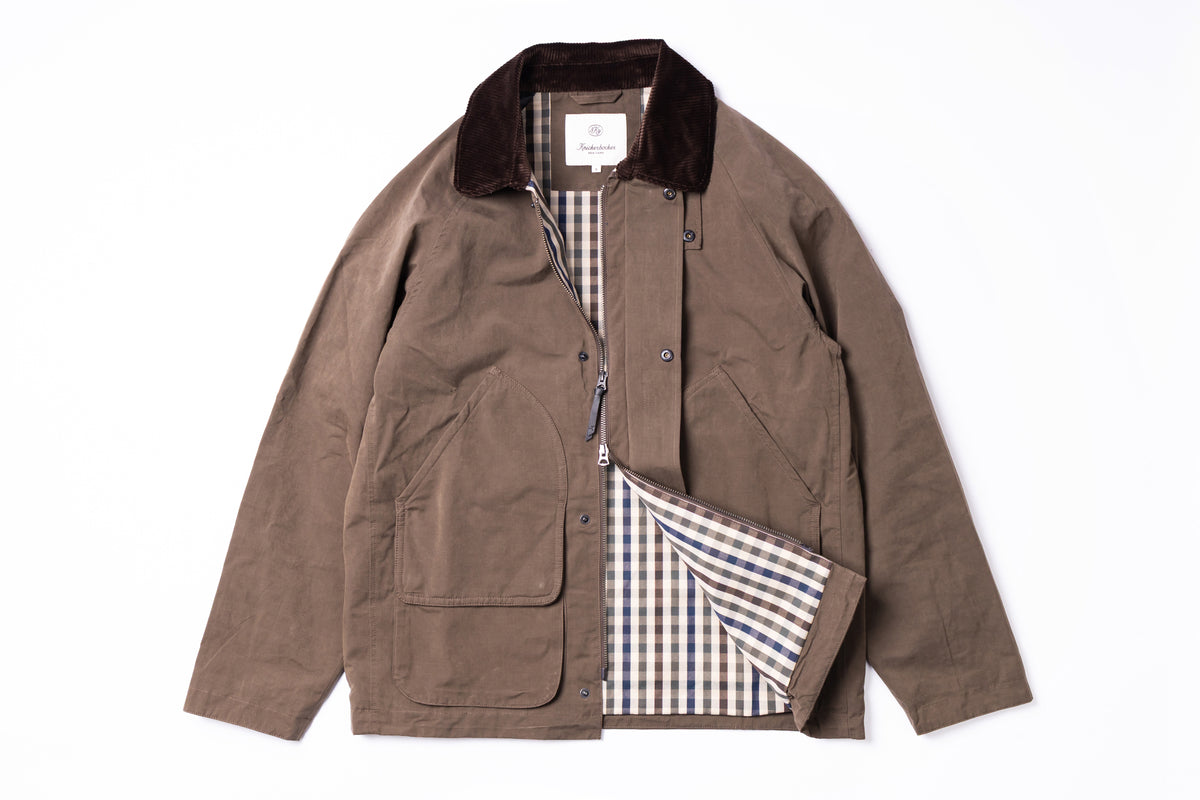 A brown waxed canvas utility jacket with a dark brown corduroy collar, featuring a plaid lining in beige, blue, and white, displayed flat against a white background.