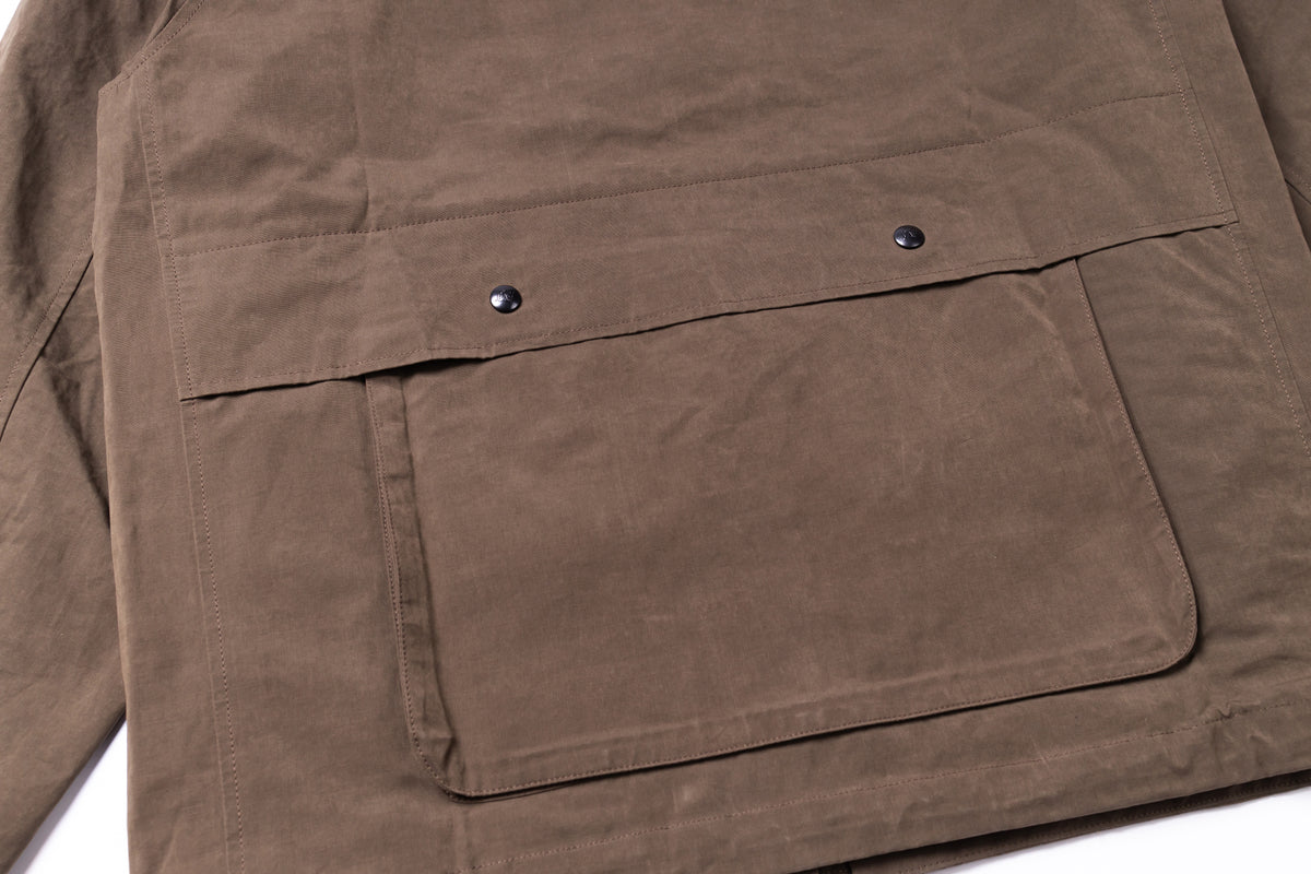 Close-up of a brown waxed canvas utility jacket showing a large front flap pocket with two metal snap buttons.