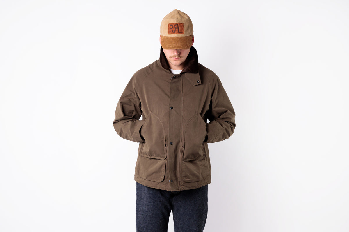 A man wearing a brown waxed canvas utility jacket with a dark brown collar, paired with dark blue jeans and a tan baseball cap, standing against a plain white background.