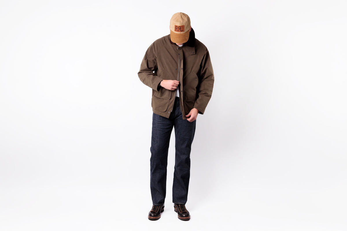 A model wearing a brown utility jacket, dark blue jeans, brown boots, and a tan baseball cap, standing against a plain white studio background.