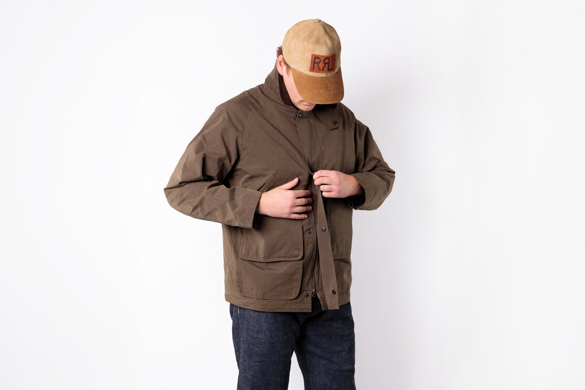 A man wearing a brown utility jacket, dark blue jeans, and a tan baseball cap with a brown logo patch, standing against a plain white background.