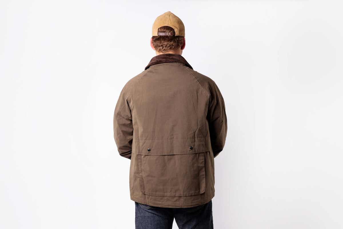 A man viewed from behind wearing a brown utility jacket with a dark brown corduroy collar, a tan baseball cap, and dark blue jeans, standing against a plain white background.
