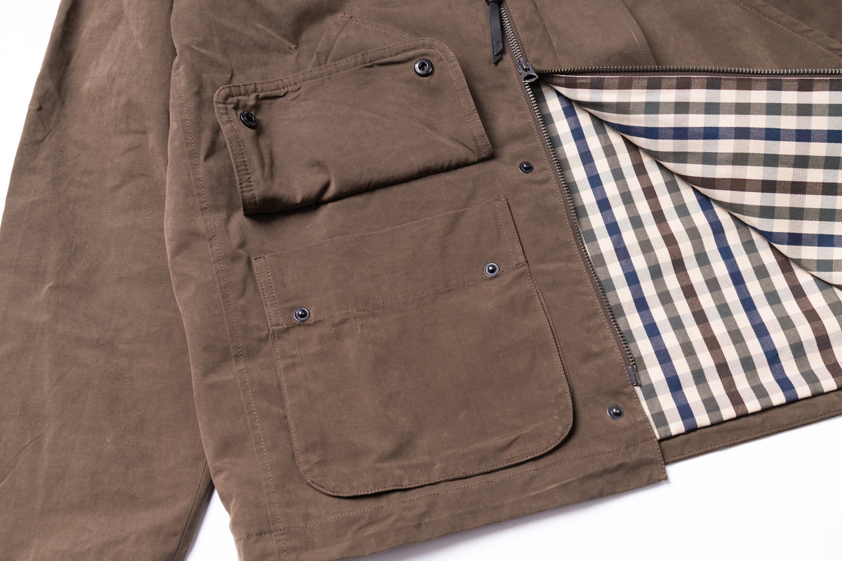 Close-up of a brown waxed canvas jacket with a zippered front, buttoned flap pockets, and a beige, blue, and brown plaid lining visible on the open side.