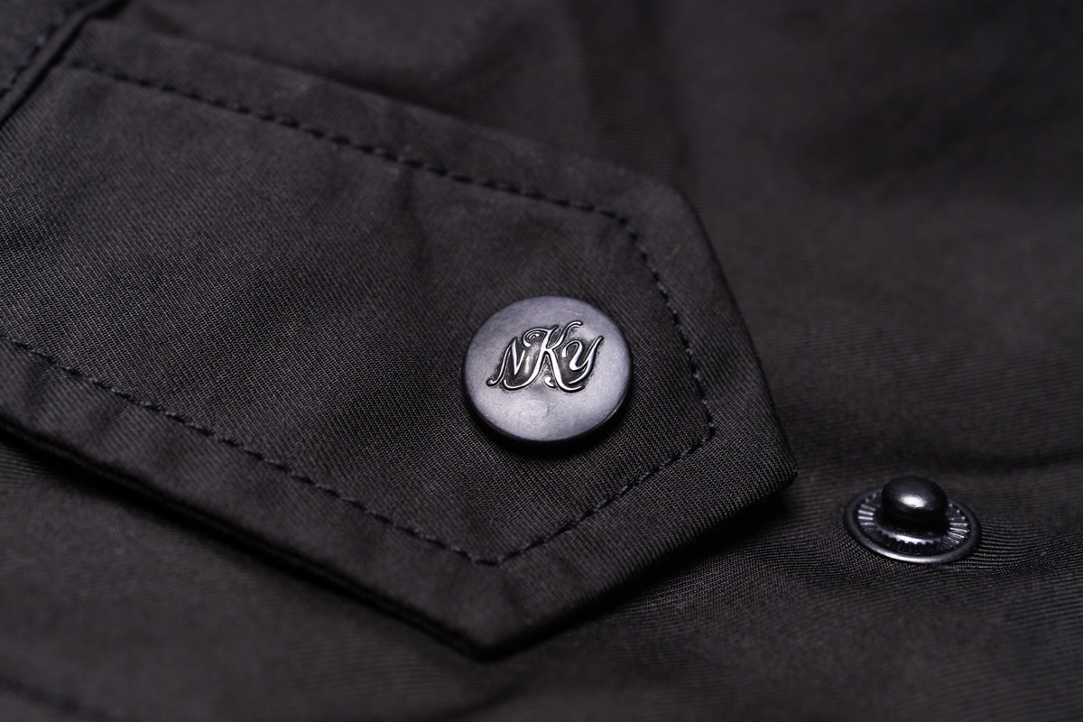 Close-up of a black jacket or overshirt fabric detail, featuring a silver-toned metal button with an engraved stylized 'BKY' logo on a pocket flap, alongside another dark metal snap.
