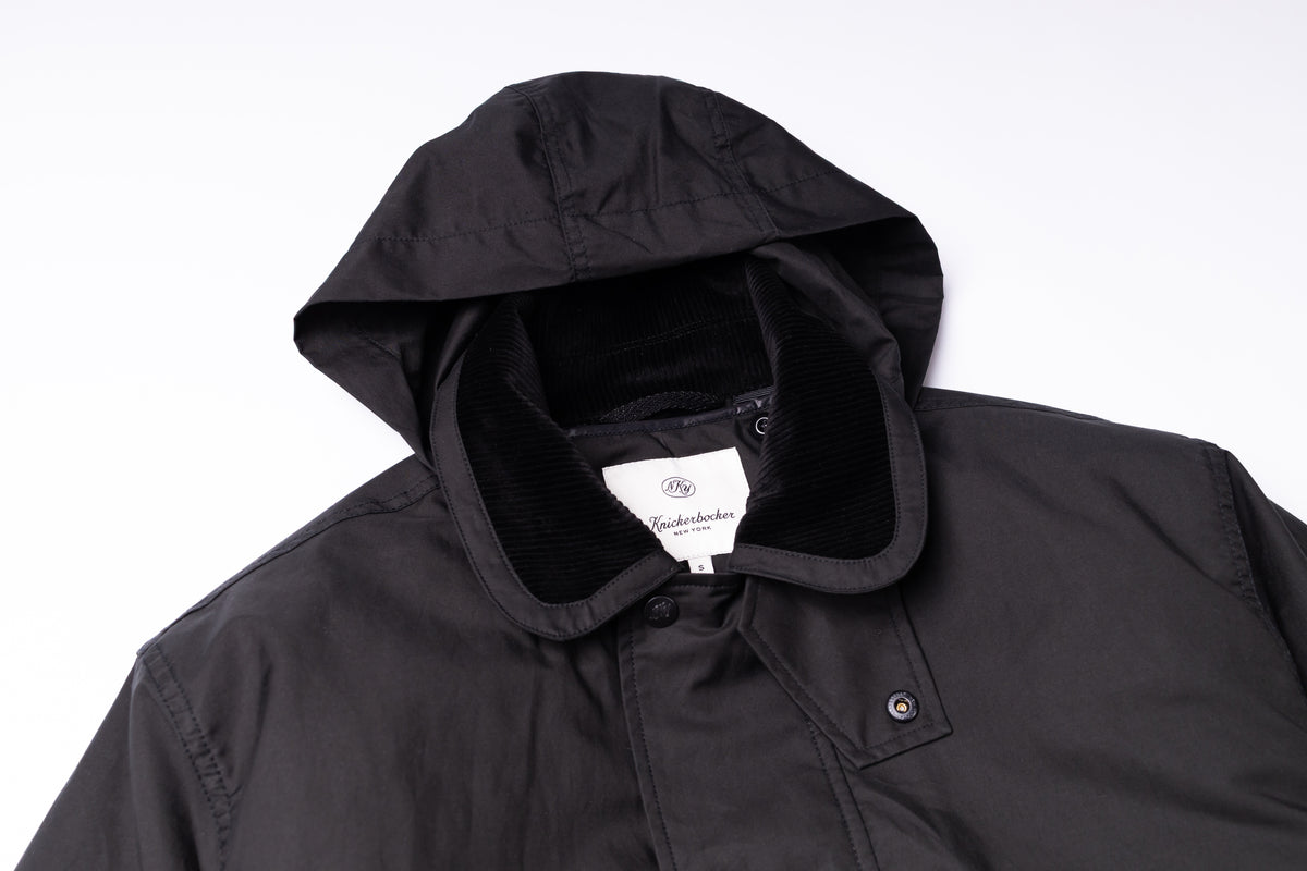 A close-up of a black hooded jacket with a black corduroy-lined hood and a white interior neck label, photographed against a plain white background.