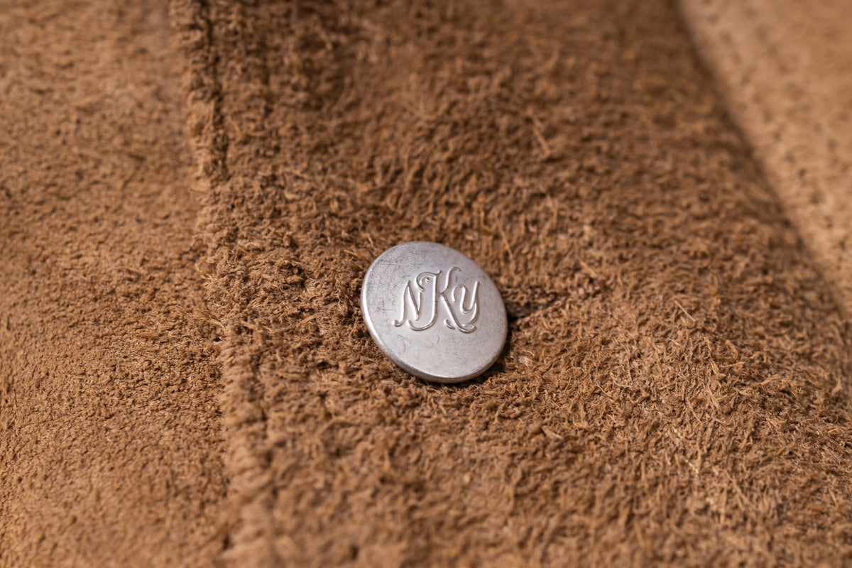 A close-up of a brown textured suede or leather surface with a small round silver button featuring the engraved initials 'NKY'.
