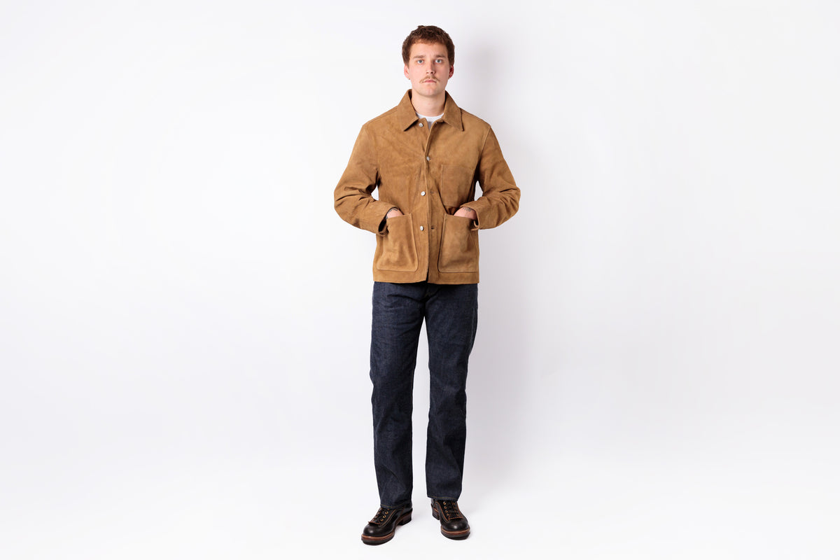 A man wearing a tan suede jacket, dark blue jeans, and brown shoes, standing against a plain white studio background.