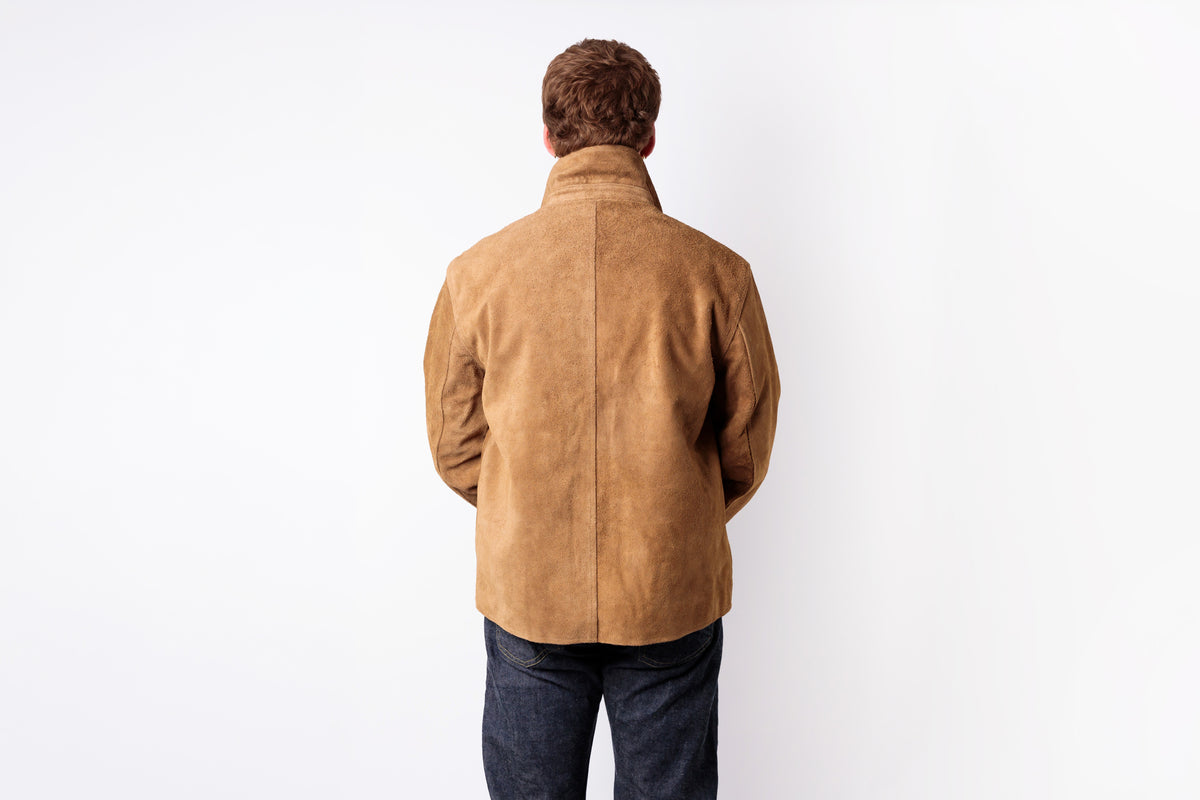 A man viewed from behind wearing a tan suede jacket and dark blue jeans, standing against a plain white background.