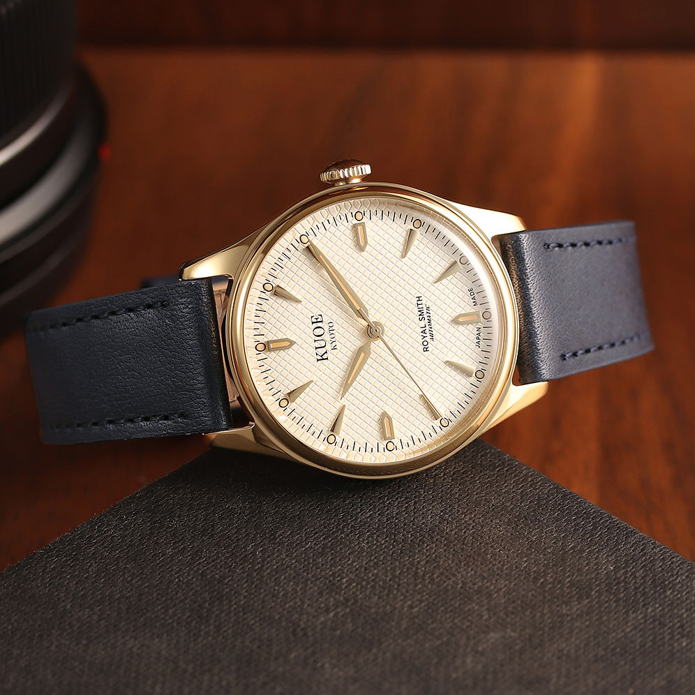A vintage-style wristwatch with a gold-tone case, cream dial, and dark blue leather strap, resting on a dark textured surface against a warm wooden background.