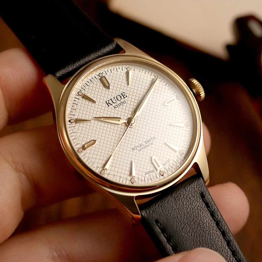 A close-up of a KUOE watch with a gold-toned case, white grid dial, and black leather strap, held in a person's hand.