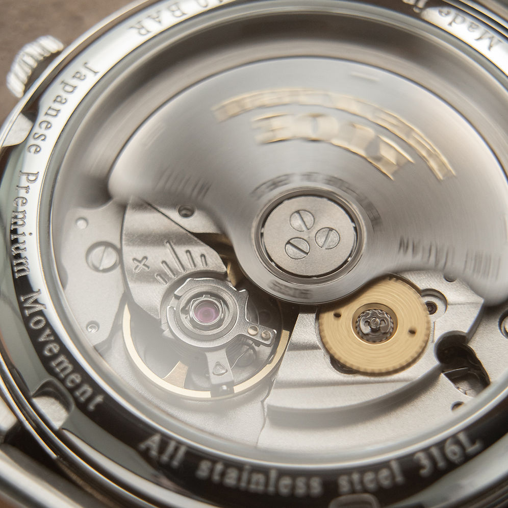 Close-up of a stainless steel watch movement with engraved text reading 'Japanese Premium Movement' and 'All stainless steel 316L', showing polished metal gears and a gold-toned wheel.