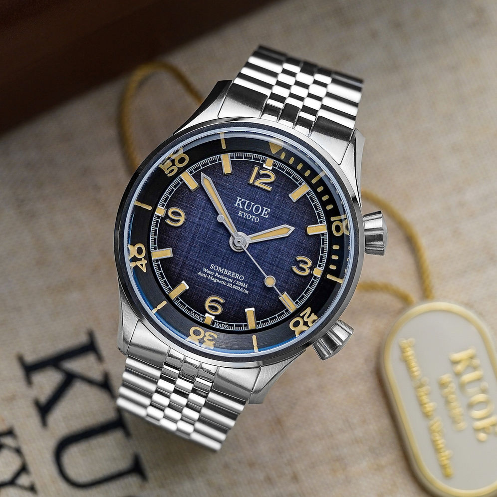 A KUOE stainless steel watch with a blue textured dial and gold-tone markers, resting on a beige textured surface next to its branded tag.