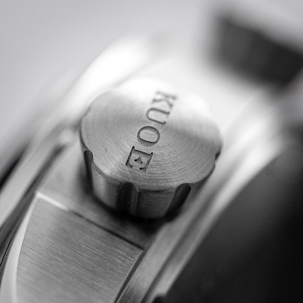 Close-up of a brushed metal watch case with a textured crown knob engraved with the brand name KUO.