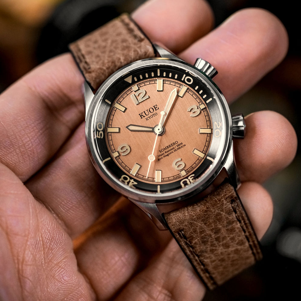 A close-up of a wristwatch held in a hand. The watch has a round stainless steel case, a warm tan dial with black Arabic numerals and hands, and a brown textured leather strap.